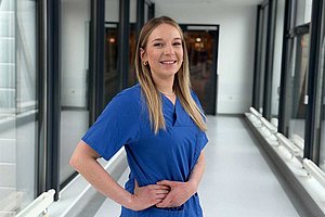 Michelle Fenzau in blauer Pflegekleidung lächelt in einem hellen Krankenhausflur in die Kamera.